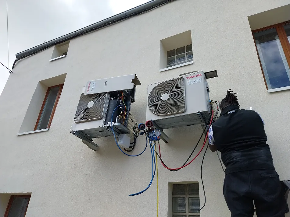 Technician wearing a black vest working on two wall-mounted Toshiba air conditioning units with gauges and colorful cables connected.