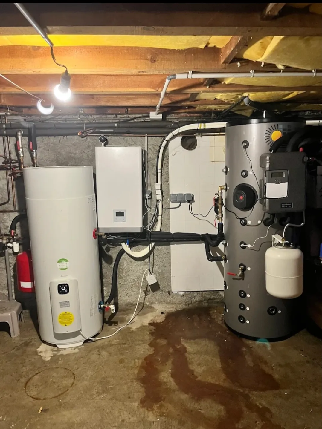 Basement utility area with water heaters, pipes, and wiring under exposed wooden ceiling beams.