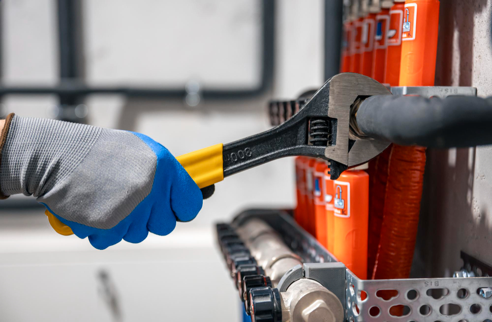 Gloved hand using adjustable wrench to tighten a pipe fitting on a manifold system with orange insulation covers.